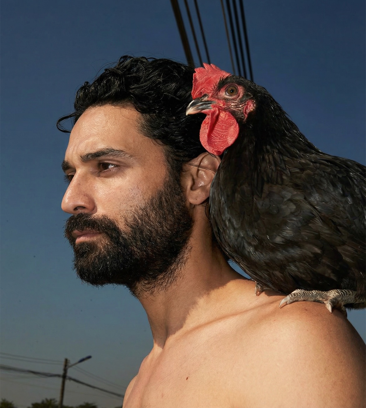 Amos Le Blanc shirtless in profile with a black rooster perched on his shoulder