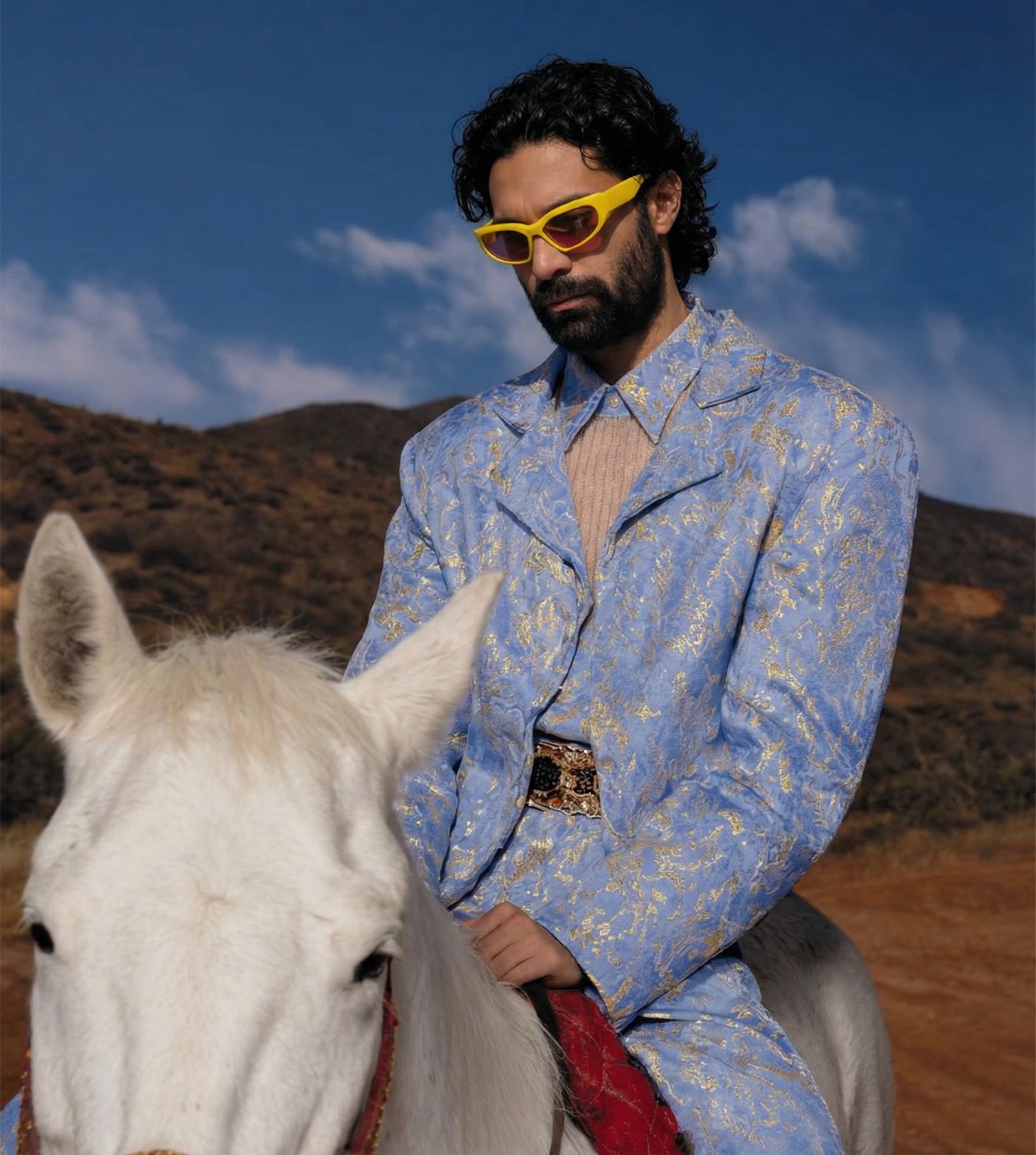 Amos Le Blanc riding a white horse in a blue and gold suit with yellow sunglasses