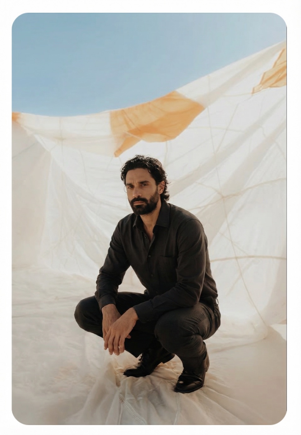 Amos Le Blanc crouching in black clothes on white draped fabric under open sky