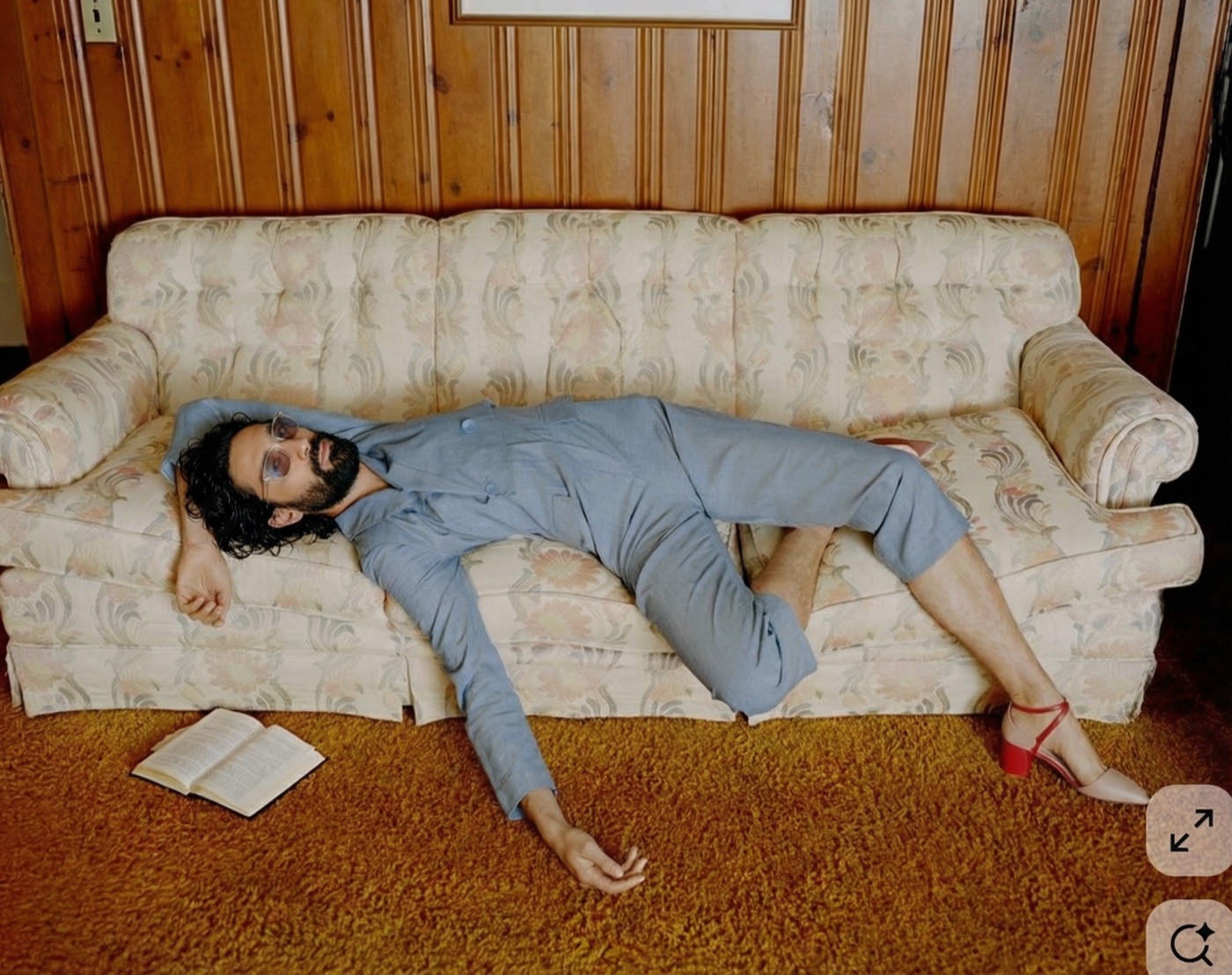 Amos Le Blanc in a grey suit sprawled across a floral vintage sofa wearing red heels