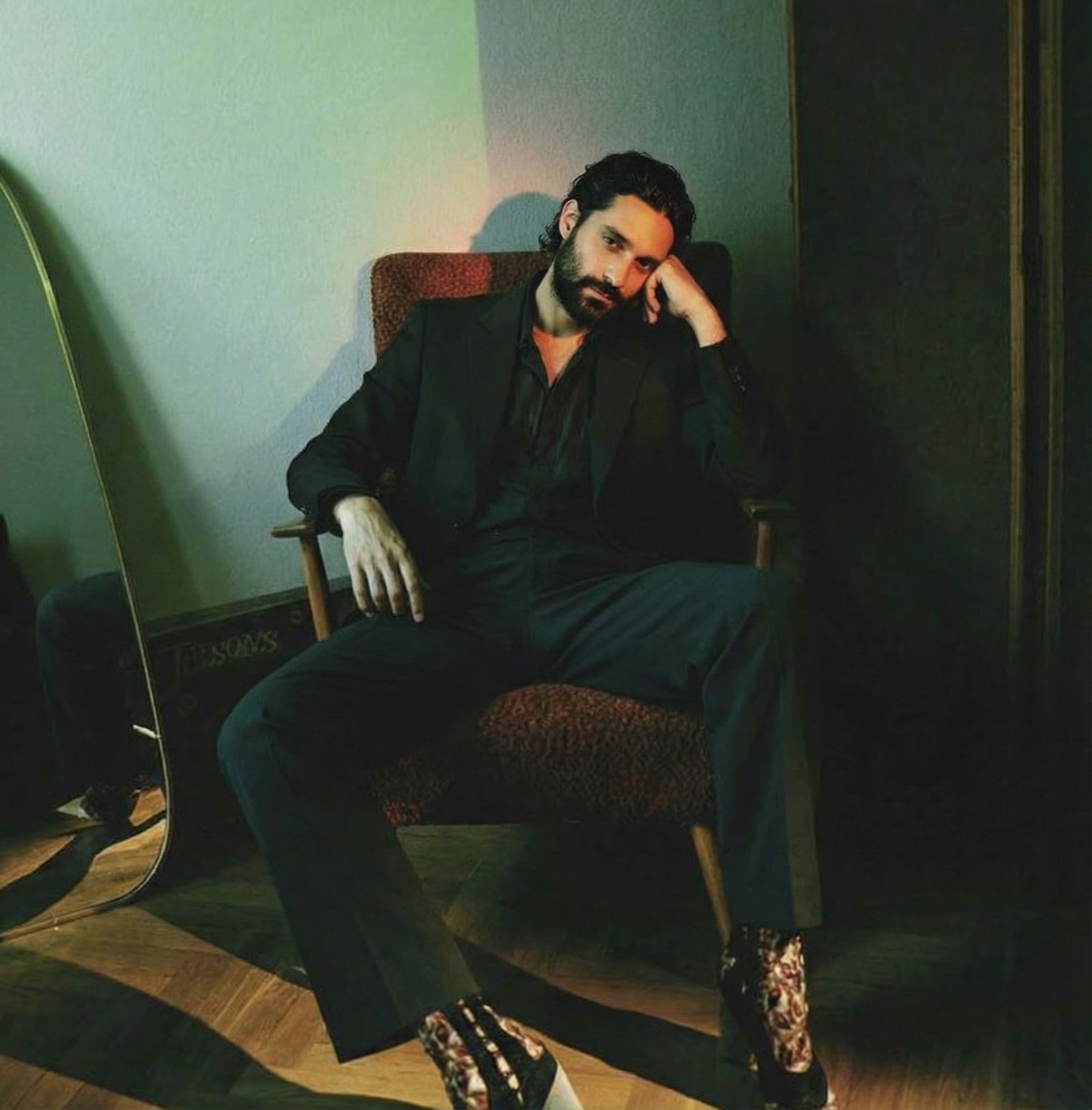 Amos Le Blanc in black seated in an armchair beside a mirror in dim green light